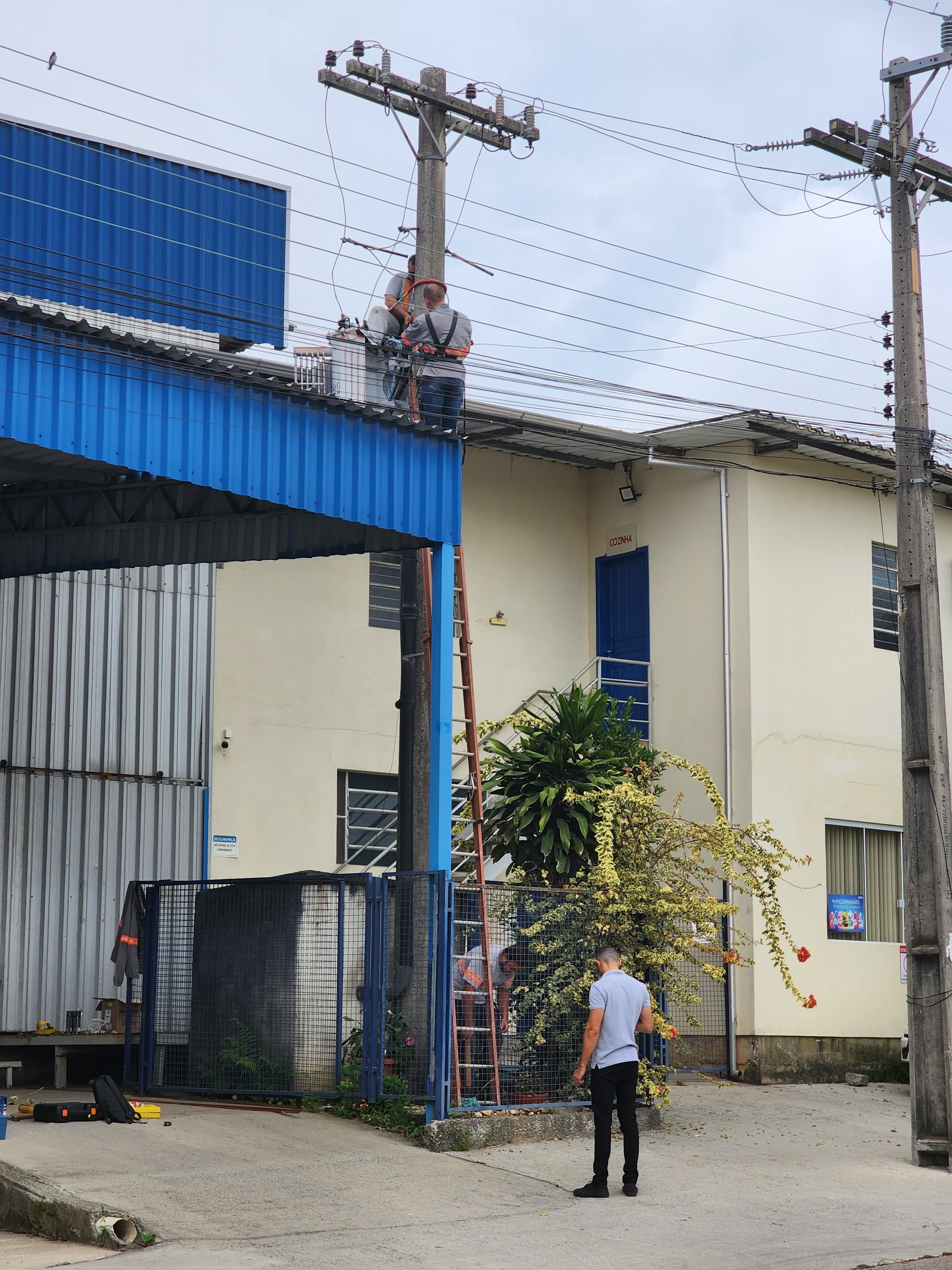 Técnico trabalhando em poste de média tensão com supervisor — serviço em altura com risco elétrico